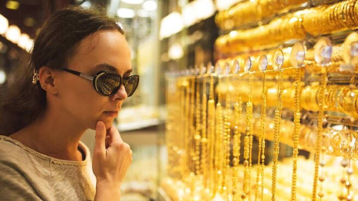 A woman wearing sunglasses closely examines gold jewellery displayed in a well-lit shop in Dubai.