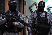 Two Hamas fighters in camouflage uniforms and black tactical gear stand armed with rifles in Lebanon. One wears a black face mask, the other a dark angular helmet mask. Urban setting with a military poster in the background.