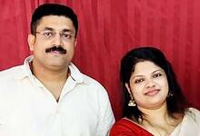 Suraj and Binsi, an Indian couple from Kerala, smiling together in a posed photo against a red curtain backdrop.