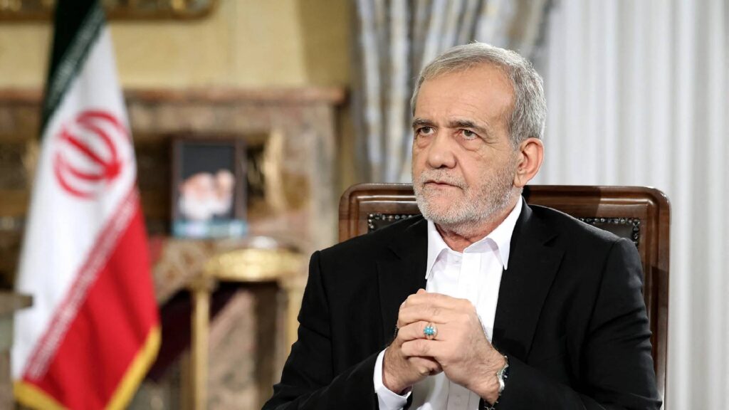 Iranian President Masoud Pezeshkian sits during a formal address, hands clasped, with the Iranian flag and a portrait in the background.
