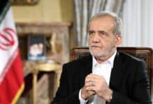 Iranian President Masoud Pezeshkian sits during a formal address, hands clasped, with the Iranian flag and a portrait in the background.