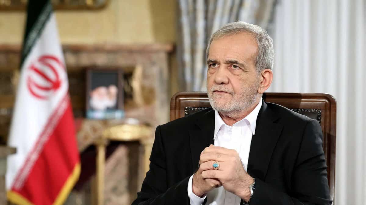 Iranian President Masoud Pezeshkian sits during a formal address, hands clasped, with the Iranian flag and a portrait in the background.