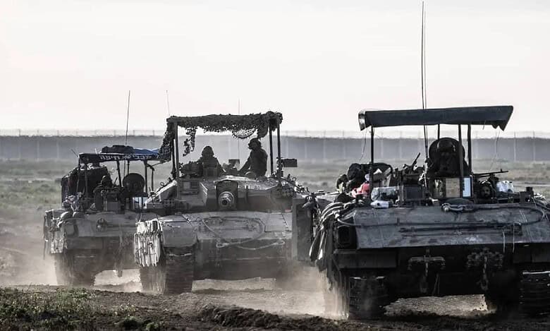 Israeli army tanks and armoured vehicles advancing through a dusty field near Gaza border during a military operation.