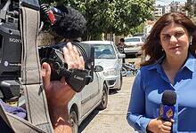 Slain Palestinian-American journalist Shireen Abu Akleh reporting from a street in the West Bank, holding an Al Jazeera microphone, with a cameraman filming her and residential buildings in the background.