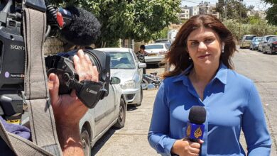 Slain Palestinian-American journalist Shireen Abu Akleh reporting from a street in the West Bank, holding an Al Jazeera microphone, with a cameraman filming her and residential buildings in the background.