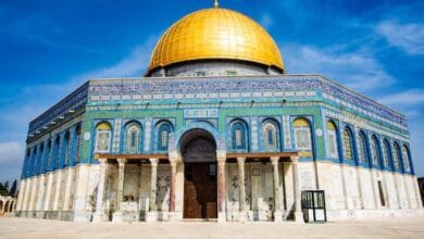 The Dome of the Rock, an Islamic shrine with a striking golden dome and intricate blue tilework, stands under a clear blue sky in Jerusalem's Al-Aqsa Mosque compound.