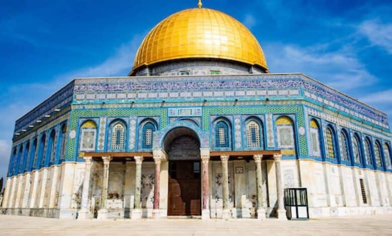 The Dome of the Rock, an Islamic shrine with a striking golden dome and intricate blue tilework, stands under a clear blue sky in Jerusalem's Al-Aqsa Mosque compound.