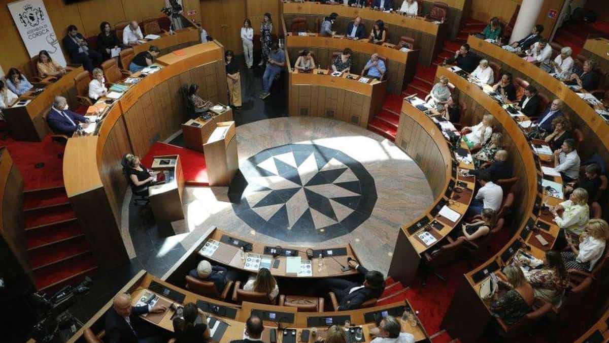 Members of the Corsican Assembly seated in a circular chamber during a formal session, with documents and microphones on desks.