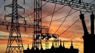 Silhouette of high-voltage transmission towers and power lines against a vibrant orange and pink sunset sky, with a power plant's cooling towers visible in the background.