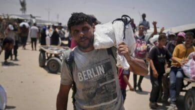 The image of 43-year-old Omar al-Hobi from Rafah carrying a bag of food over his shoulder, wearing a "Berlin Munich" t-shirt, walks through a crowded area with tents and carts in the background in Gaza.