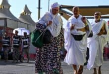 Three pilgrims in ihram walk on a road in Mina during Haj 2025, with tents, security personnel, and a bus in the background under a clear blue sky.