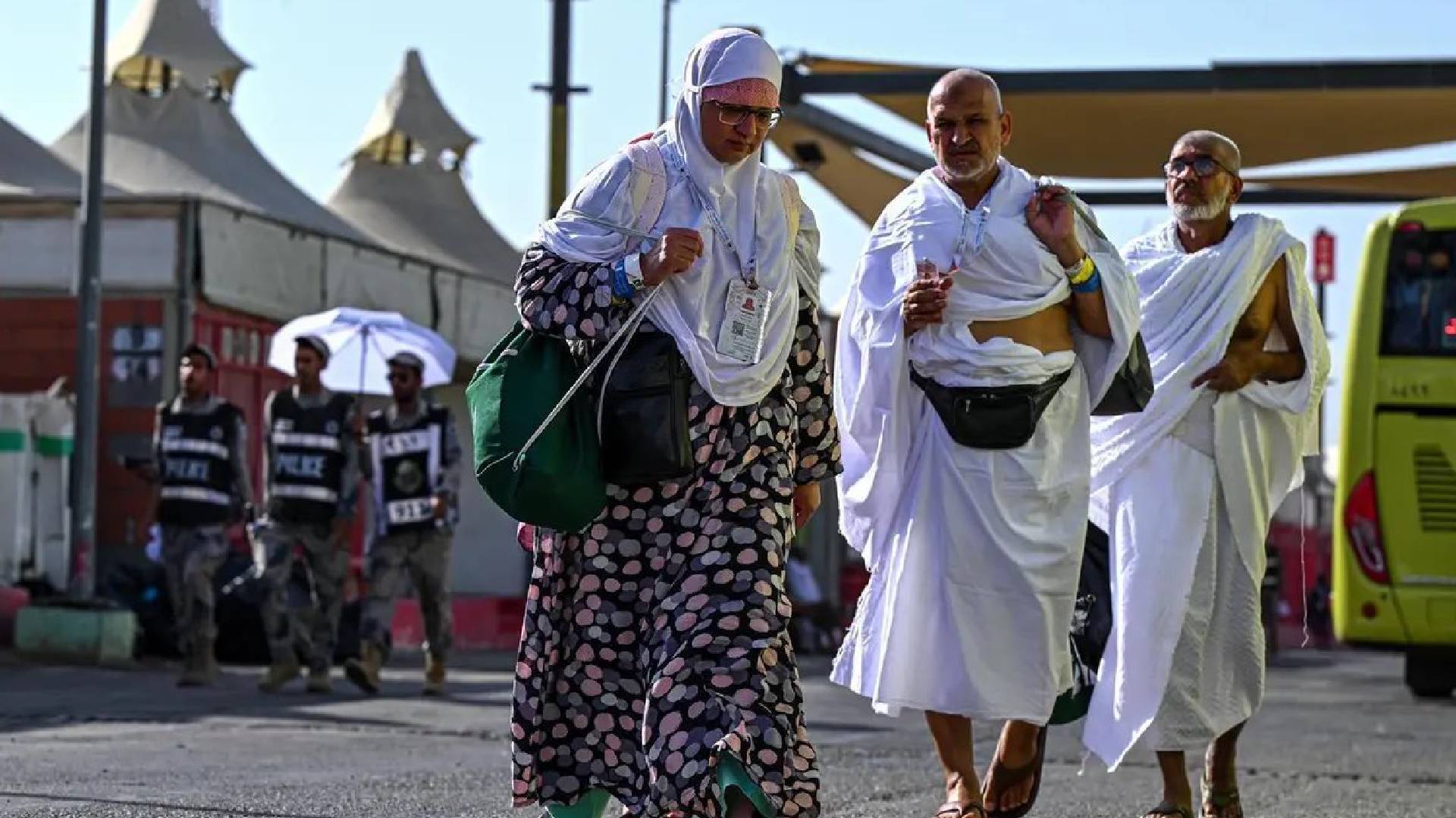 Three pilgrims in ihram walk on a road in Mina during Haj 2025, with tents, security personnel, and a bus in the background under a clear blue sky.
