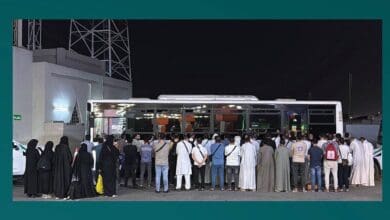 The image displays a group of 53 expatriates stands in front of a bus at night in Makkah, Saudi Arabia, after being arrested by Haj Security Forces for attempting to perform Haj without a permit on June 5.