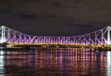 A night view of the Howrah Bridge in Kolkata, India, illuminated with purple lights and reflecting beautifully on the Hooghly River.