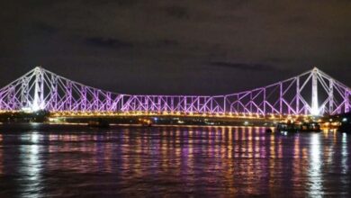 A night view of the Howrah Bridge in Kolkata, India, illuminated with purple lights and reflecting beautifully on the Hooghly River.