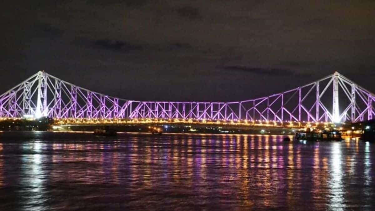 A night view of the Howrah Bridge in Kolkata, India, illuminated with purple lights and reflecting beautifully on the Hooghly River.