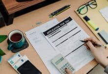 The image of a person’s hand holding a pen fills out a tax application form on a wooden desk, alongside an invoice, a calculator displaying £65, a cup of tea, euro banknotes, glasses, pencils, a highlighter, and a potted plant.