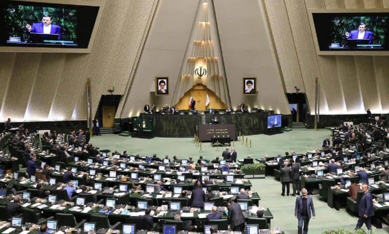 The image of a Iranian parliamentary chamber with lawmakers at computer desks, central podium with emblem and portraits, and screens showing a speaker, under a triangular ceiling, June 25, 2025.