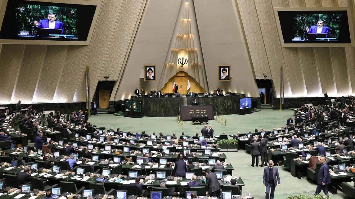 The image of a Iranian parliamentary chamber with lawmakers at computer desks, central podium with emblem and portraits, and screens showing a speaker, under a triangular ceiling, June 25, 2025.