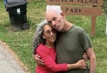 Weinstein, 70, and Haggai, 72, both Israeli-American hostages, embrace in front of a sign reading "Bayview Village Park." Weinstein, in a red shirt, has curly gray hair, and Haggai, in a green shirt, has white hair. A black trash bin and a notice about coyotes are in the background. They stand on a grassy area near a paved path.