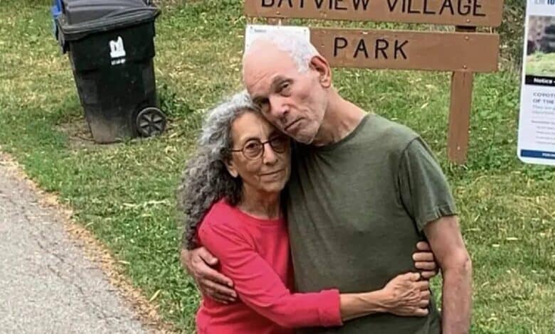 Weinstein, 70, and Haggai, 72, both Israeli-American hostages, embrace in front of a sign reading "Bayview Village Park." Weinstein, in a red shirt, has curly gray hair, and Haggai, in a green shirt, has white hair. A black trash bin and a notice about coyotes are in the background. They stand on a grassy area near a paved path.