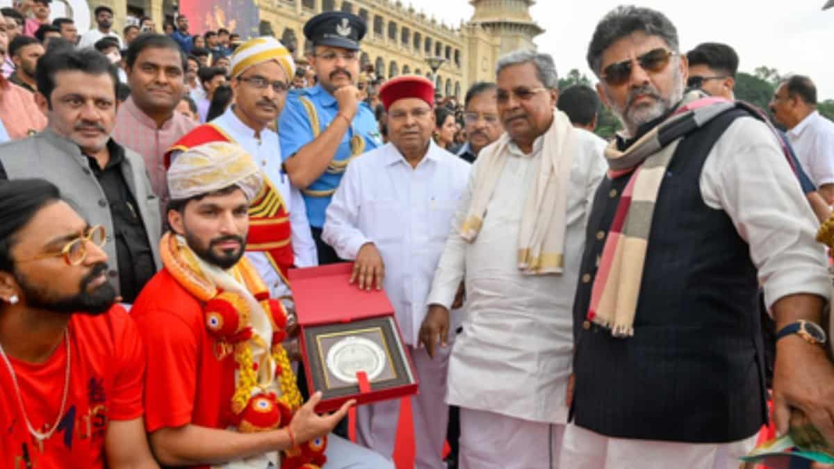 Group photo of Karnataka CM Siddaramaiah, DyCM DK Shivakumar, and Governor Thaawarchand Gehlot with a young man in traditional attire holding an award. They stand on a decorated stage with a historic building backdrop, surrounded by officials and crowd, celebrating an event.