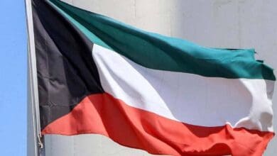 The national flag of Kuwait waving in the wind, showing horizontal stripes of green, white, and red with a black trapezoid on the hoist side, against a light-coloured background.