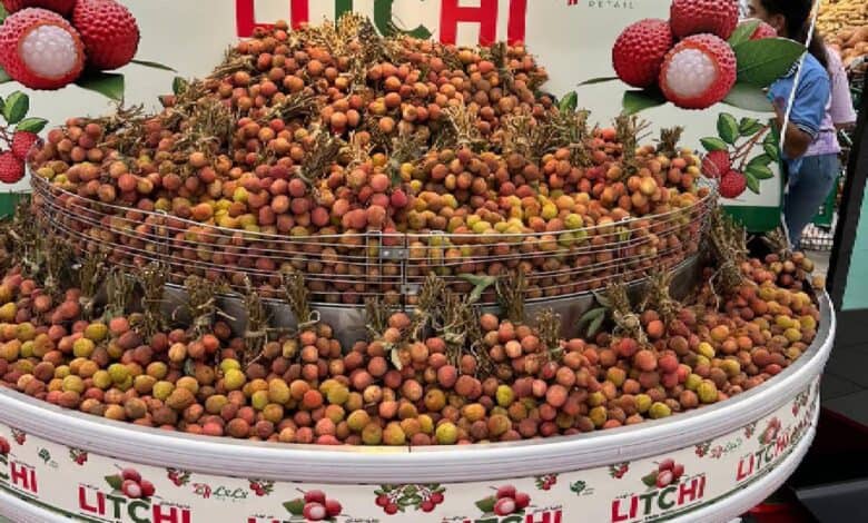 A large display of fresh litchi fruits in a wire basket at a market, with a backdrop featuring images of litchi and the word "LITCHI" in bold red letters. A person is partially visible shopping nearby.