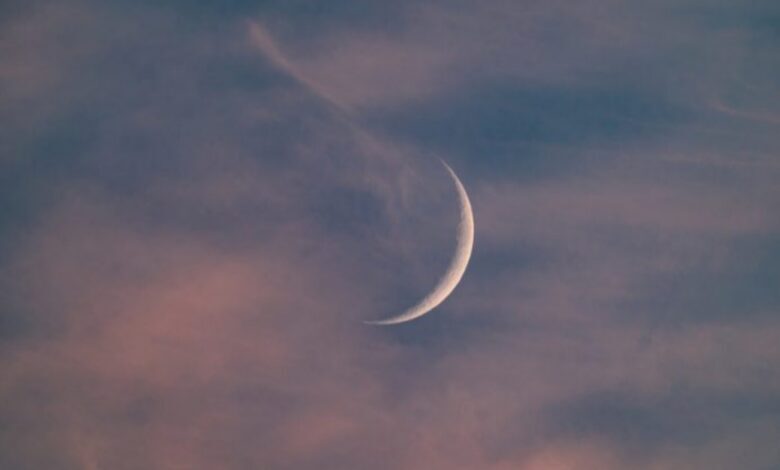 The image of crescent moon glows in a cloudy sky at dusk, with shades of pink, purple, and blue blending across the horizon. Wispy clouds are faintly illuminated, creating a serene and tranquil atmosphere.