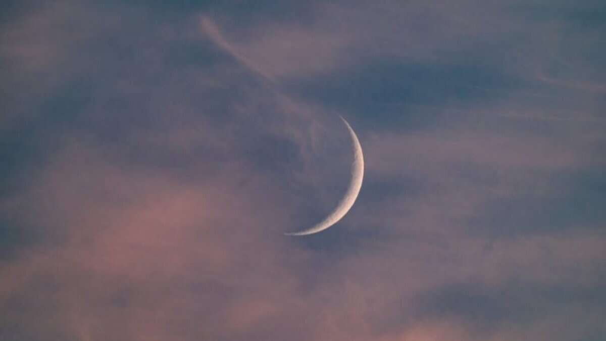 The image of crescent moon glows in a cloudy sky at dusk, with shades of pink, purple, and blue blending across the horizon. Wispy clouds are faintly illuminated, creating a serene and tranquil atmosphere.