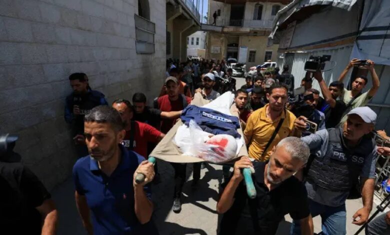 The image of a group of mourners, including men in casual clothing, carry a stretcher with the body of Palestinian journalist Ismail Baddah, draped in a white cloth and a blue jacket, through a narrow street in Gaza City.