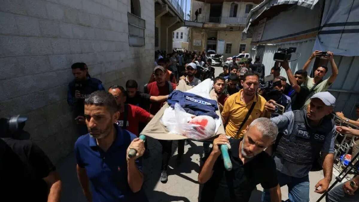 The image of a group of mourners, including men in casual clothing, carry a stretcher with the body of Palestinian journalist Ismail Baddah, draped in a white cloth and a blue jacket, through a narrow street in Gaza City.