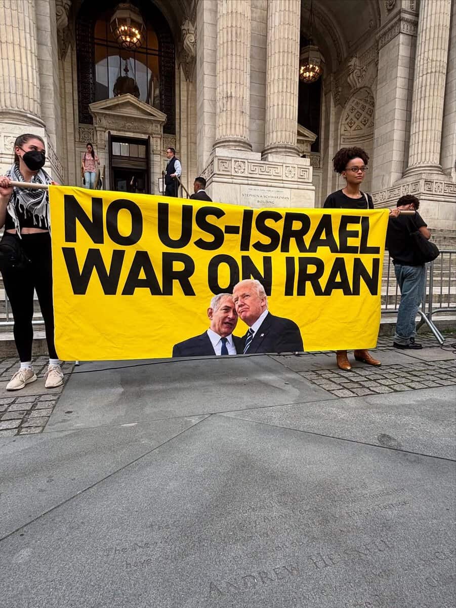 Two protesters hold a large yellow banner outside the New York Public Library reading “NO US-ISRAEL WAR ON IRAN,” with images of Joe Biden and Benjamin Netanyahu. The library’s grand pillars and entrance are visible in the background.