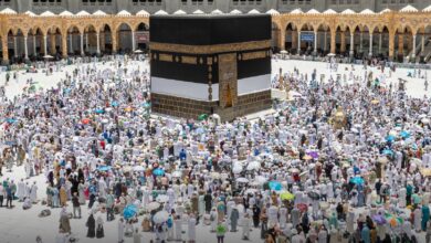 The image of a large crowd of pilgrims gathers around the Kaaba at the Grand Mosque in Makkah, many holding umbrellas to shield from the sun. The black and gold-draped Kaaba stands at the centre of the courtyard, surrounded by white-clad worshippers performing rituals.