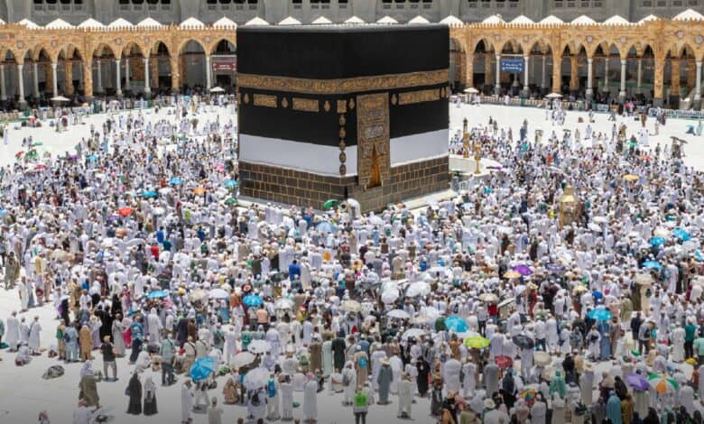 The image of a large crowd of pilgrims gathers around the Kaaba at the Grand Mosque in Makkah, many holding umbrellas to shield from the sun. The black and gold-draped Kaaba stands at the centre of the courtyard, surrounded by white-clad worshippers performing rituals.
