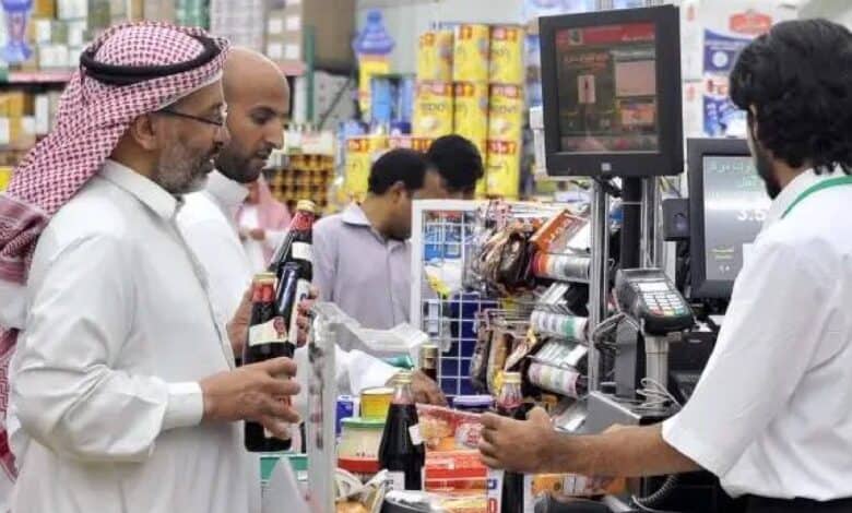 The image of a customers in traditional Saudi attire queue at a supermarket checkout counter, purchasing groceries and bottled beverages, with a cashier scanning items.