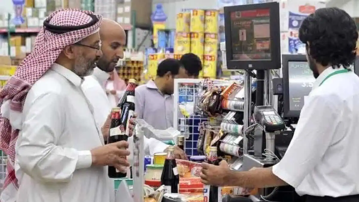 The image of a customers in traditional Saudi attire queue at a supermarket checkout counter, purchasing groceries and bottled beverages, with a cashier scanning items.