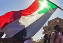 A group of people in Sudan walk under a large waving Sudanese flag, with sunlight shining through its green, white, black, and red colours. A brick building is visible in the background under a clear blue sky.