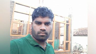 The image of a man in a green shirt stands in front of a partially constructed brick building at a rural construction site.