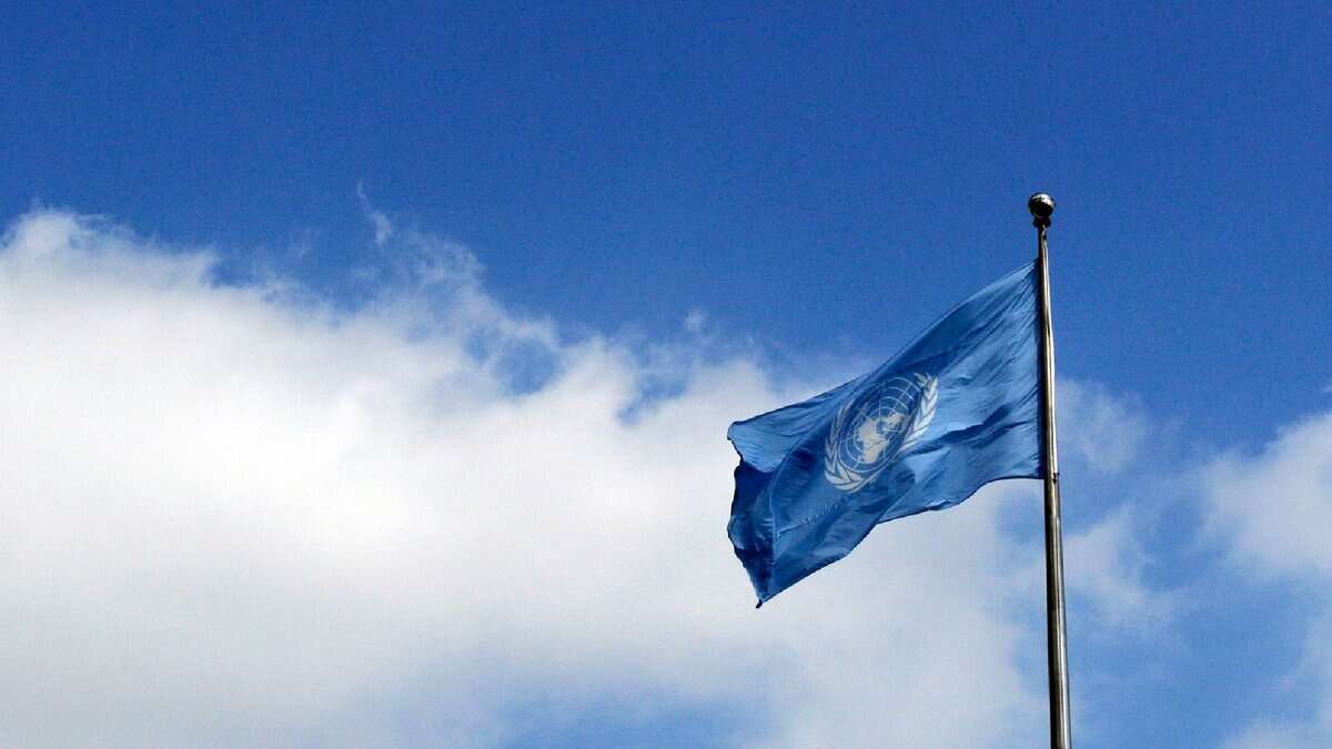 The image of United Nations flag waving against a bright blue sky with scattered clouds.