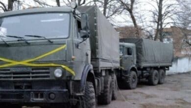 A line of olive‑drab military cargo trucks with canvas-covered beds, parked in a wooded yard — the lead vehicle marked with a prominent yellow “X.” Sources