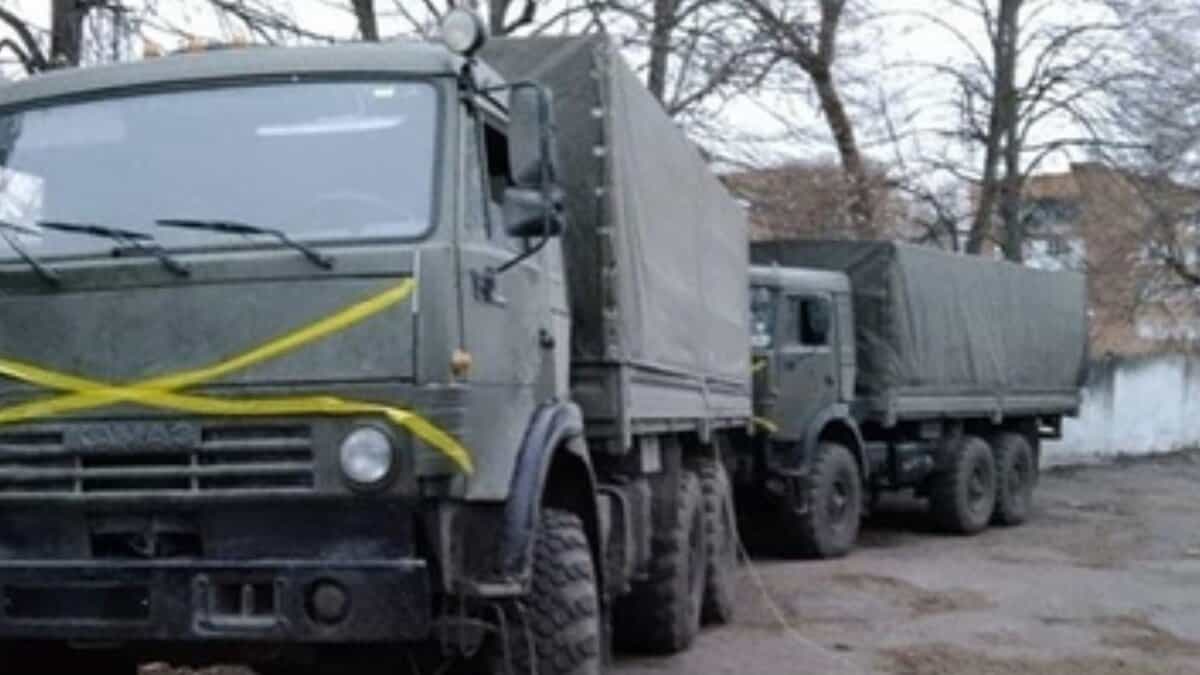 A line of olive‑drab military cargo trucks with canvas-covered beds, parked in a wooded yard — the lead vehicle marked with a prominent yellow “X.” Sources