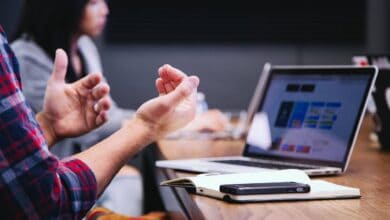 The image of a workplace meeting with a person gesturing beside a laptop, notebook, and mobile phone on a conference table.