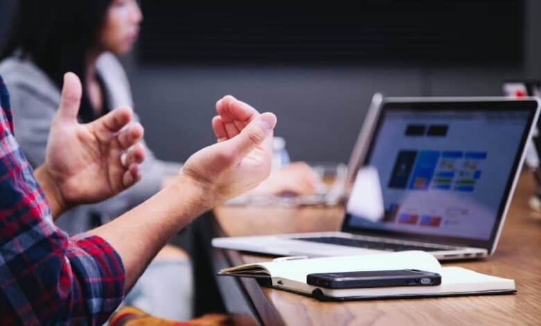 The image of a workplace meeting with a person gesturing beside a laptop, notebook, and mobile phone on a conference table.