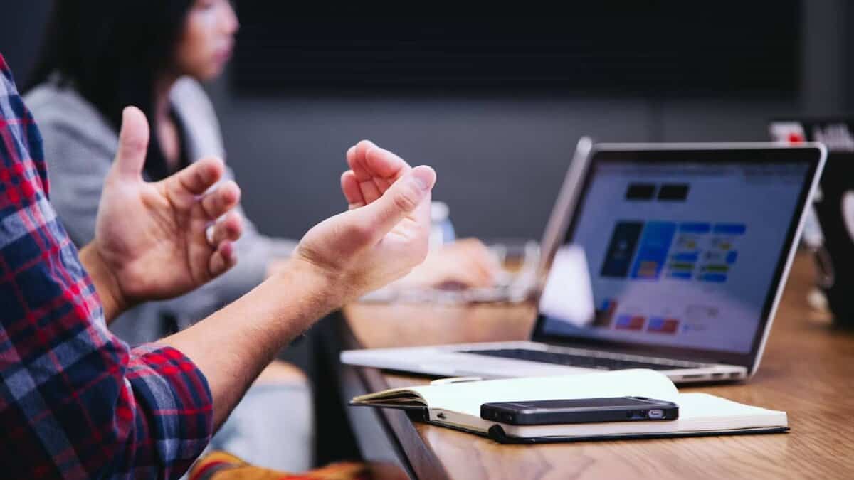 The image of a workplace meeting with a person gesturing beside a laptop, notebook, and mobile phone on a conference table.