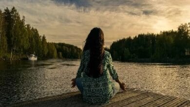 A person with long hair sits cross-legged on a wooden dock by a calm lake, meditating at sunset. Trees line the shore, and a boat is visible in the distance. meditation techniques yoga practices more descriptive