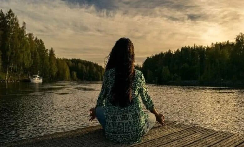 A person with long hair sits cross-legged on a wooden dock by a calm lake, meditating at sunset. Trees line the shore, and a boat is visible in the distance. meditation techniques yoga practices more descriptive
