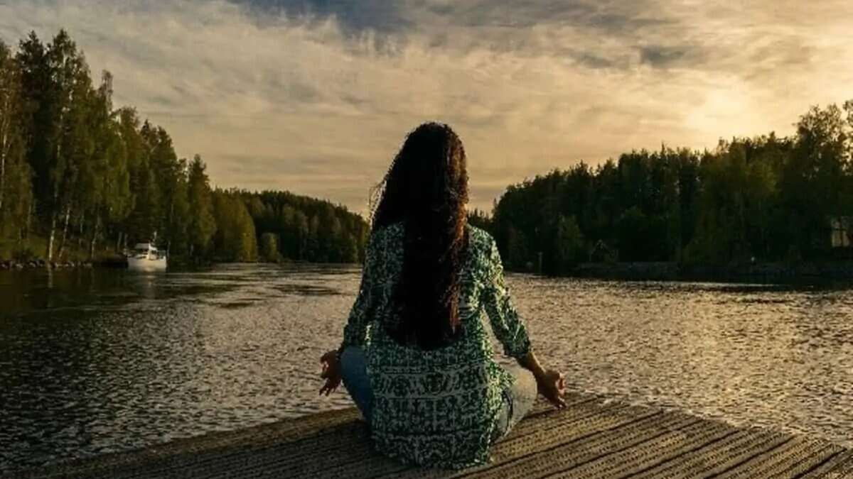 A person with long hair sits cross-legged on a wooden dock by a calm lake, meditating at sunset. Trees line the shore, and a boat is visible in the distance. meditation techniques yoga practices more descriptive