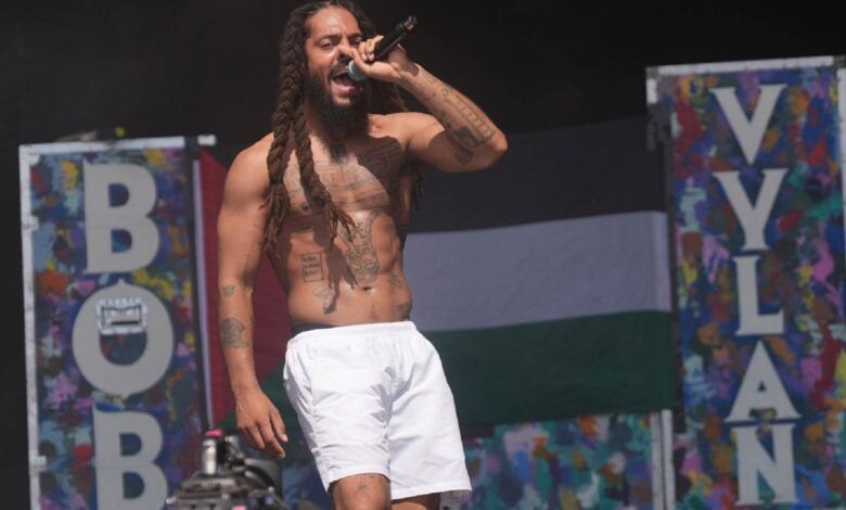 Shirtless rapper Bob Vylan performs on stage at Glastonbury Festival, with a Palestinian flag and colourful set decorations in the background.