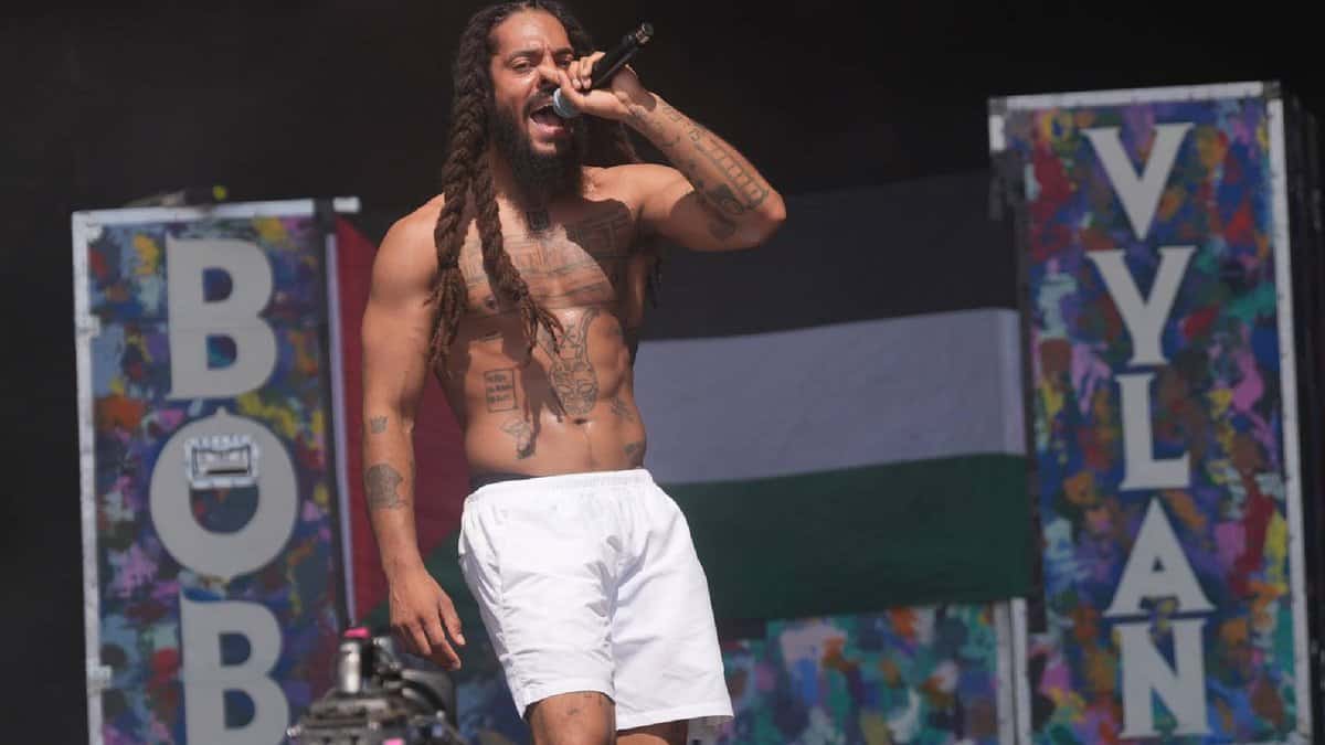 Shirtless rapper Bob Vylan performs on stage at Glastonbury Festival, with a Palestinian flag and colourful set decorations in the background.